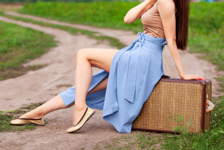 Pretty girl is sitting on an old suitcase on a country roadの写真素材