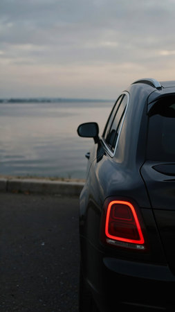 Model Version: pro | Model Category: Realism | Prompt: Photorealistic sleek black luxury car, rear view, parked near a calm body of water at dusk, vivid red taillight glow, glossy black paint, subtle ambient light reflections, visible side mirror with soft highlights, softly blurred background, serene horizon, muted twilight sky tones, still water, moody cinematic atmosphere, elegant composition, tranquil and sophisticated, dramatic lighting, shallow depth of field, professional automotive photography, premium advertising quality, 4K. | Enhanced Prompt: A photorealistic, sleek black luxury car, captured from a precise rear view, is parked elegantly near a perfectly calm body of water at the moment of dusk. The vehicle's glossy black paint reflects the subtle ambient light and the vivid, intense red glow emanating from its taillights. Visible in sharp detail is the side mirror, catching soft, elongated highlights. The background is softly blurred, emphasizing the car, revealing a serene horizon line where muted twilight sky tones meet the still water. The composition is elegant and tranquil, utilizing dramatic lighting and a shallow depth of field to create a moody, sophisticated, cinematic atmosphere. Professional automotive photography, premium advertising quality, 4K.の素材