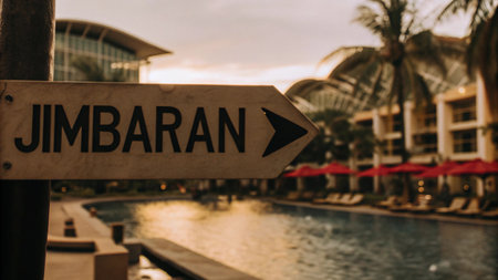 A clear sign guides visitors to Jimbaran, Bali, featuring a beautiful resort pool and swaying palm trees under a sunny sky. Find your way to this idyllic tropical paradise for a relaxing getaway.の素材