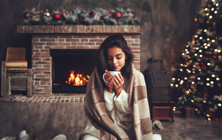 Young woman in a warm sweater with a cup of hot drink by the fireplace at Christmas.の素材
