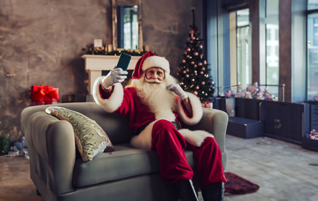 Santa Claus is sitting on the sofa and making selfie with his smartphone.の素材