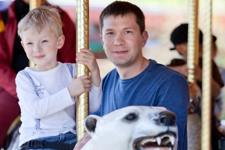 excited little boy riding a merry-go-round at the amusement park with his fatherの写真素材