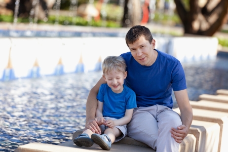 family of two enjoying time together outdoors, young handsome father and his adorable son laughing with his eyes shut sitting by the fountainの写真素材