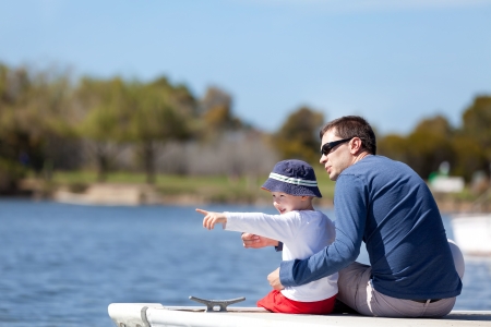 young father and his son sitting at the marina dock and spending fun time together; adorable little boy pointing with his finger and showing something to his fatherの写真素材