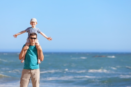 little smiling boy sitting at his young father shoulders and enjoying summer time at the beachの写真素材