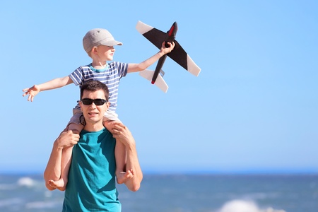 smiling positive young man and his excited son sitting on father's shoulders and playing a toy plane at the beachの写真素材