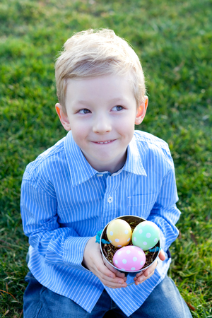cute smiling boy holding bucket with colorful eggs sitting on grass at easter timeの写真素材