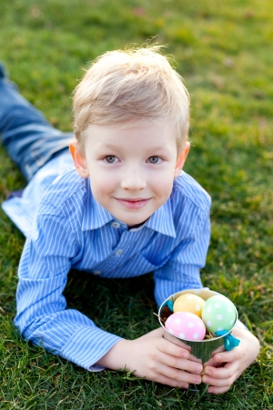 cute smiling boy holding bucket with colorful eggs lying on grass at easter timeの写真素材