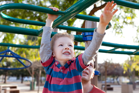 little positive boy at the monkey bars and his father watching and helping