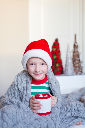 cute smiling boy sitting cozy in the blanket at home, holding cup of hot cocoa at christmas timeの写真素材