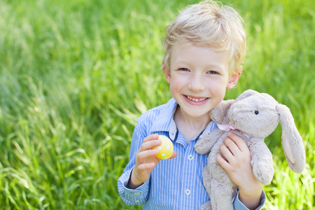 smiling cheerful little boy holding colorful easter egg and bunny toy at spring time in the park