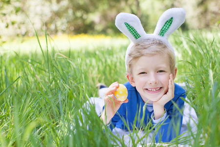 little smiling boy with bunny ears holding easter egg after egg hunt in the park lying in the grass