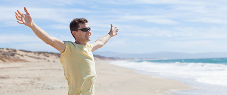 young smiling man with arms wide open at the beach enjoying the moment of lifeの写真素材
