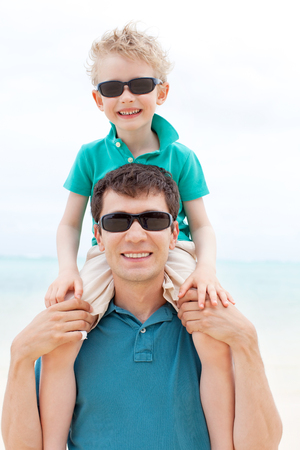 smiling little boy sitting on his father shoulders enjoying summer vacation at the beachの写真素材