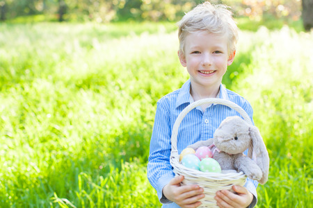 smiling cheerful boy holding basket with colorful easter eggs and bunny in the park enjoying spring...