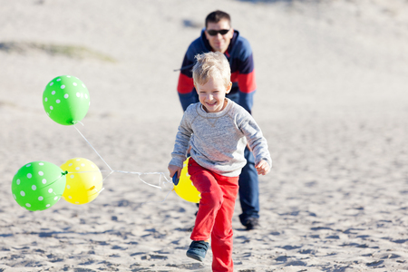 little excited boy running at the beach with colorful balloons with his father watchingの写真素材