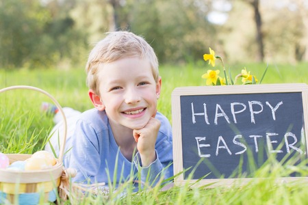 portrait of smiling boy after easter egg hunt with basket full of colorful eggs and blackboard with text "happy easter!"の写真素材