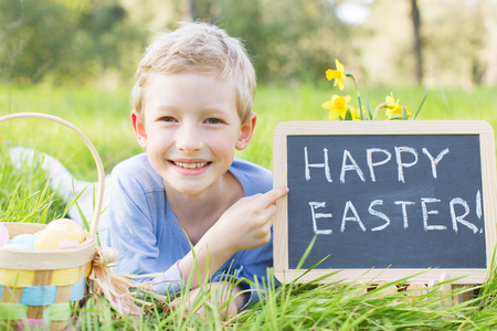 portrait of smiling boy after easter egg hunt with basket full of colorful eggs and blackboard with text "happy easter!"の写真素材