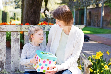 young beautiful mother and her adorable son sitting together in the park at spring time celebrating mother's day, boy giving her mother present and looking at each otherの写真素材