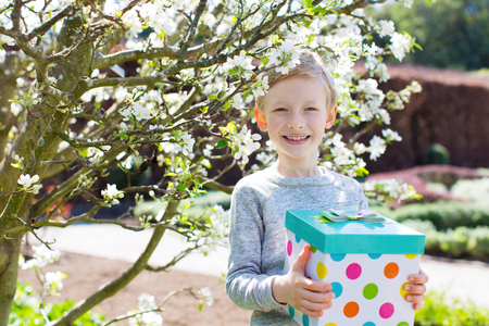 cheerful smiling boy holding colorful present for mother's day or other celebration by the blooming tree at park at spring timeの写真素材