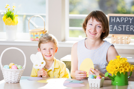 happy smiling family of two enjoying easter time at home, doing crafts, little boy holding easter bunny shaped candy, easter themed decorations and blackboard sign "happy easter!" in the backgroundの写真素材