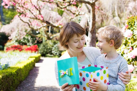 young beautiful mother and her adorable son sitting together in the park at spring time celebrating mother's day, boy giving her mother present, mother opening it and looking happyの写真素材