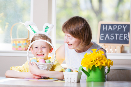 happy smiling family of two enjoying easter time at home, doing crafts, little boy with bunny ears holding basket with colorful easter eggs, easter themed decorations and blackboard sign "happy easter!" in the backgroundの写真素材
