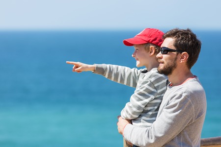 young father with a beard and his little son pointing at something with finger with beautiful blue ocean in the background enjoying time togetherの写真素材
