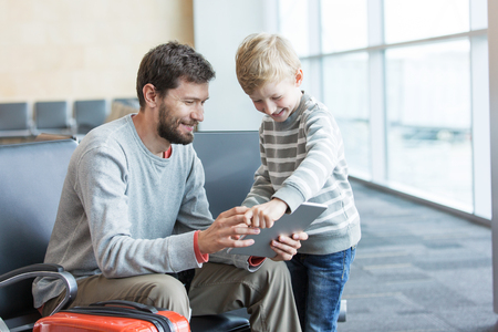 happy positive family of two, father with a beard and little son, at the airport waiting for departure and playing at tablet, travel and technology conceptの写真素材