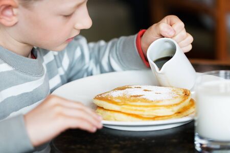beautiful little boy pouring maple syrup at pancakes for breakfastの写真素材