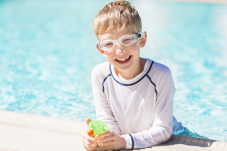 smiling excited boy with beach toy in goggles and rashguard enjoying warm weather in swimming pool, vacation and sun protection conceptの写真素材