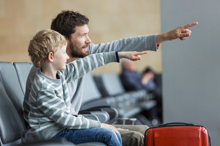 family of two sitting waiting for airplane departure and pointing together at something at airport terminal, vacation and transportation conceptの写真素材