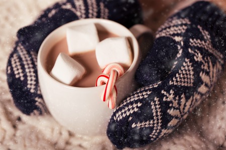 hands in cozy mittens holding hot chocolate in the cup with marshmallows and candy canes in snowy weather, christmas and winter time conceptの写真素材