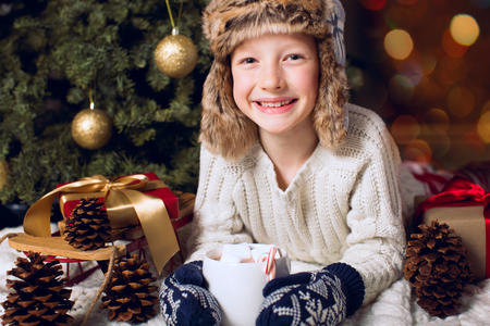 smiling cheerful little boy in cozy hat holding cup with hot chocolate at christmas time by the tree with presents, christmas and holiday conceptの写真素材