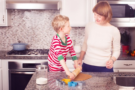 cheerful kid rolling dough for gingerbread cookies, family of mother and son enjoying christmas time at homeの写真素材