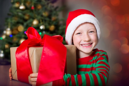 smiling happy boy in elf hat holding huge christmas present by the tree waiting for a miracle at holiday timeの写真素材