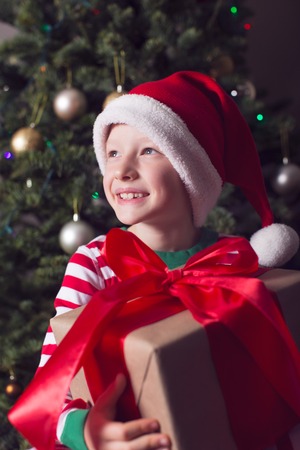 smiling happy boy in elf hat holding huge christmas present by the tree waiting for a miracle at holiday timeの写真素材