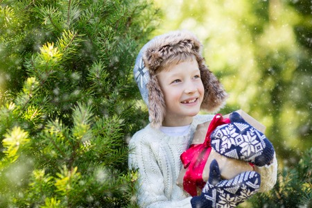 beautiful smiling boy in trapper hat, sweater and mittens holding nicely wrapped christmas gift by the tree or in the forest enjoying snowy cold winter weather, holiday or happiness conceptの写真素材