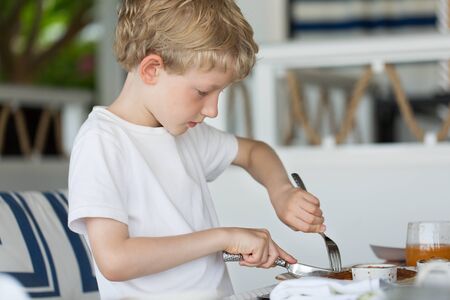 little serious boy eating breakfast with fork and knife at hotel or resort