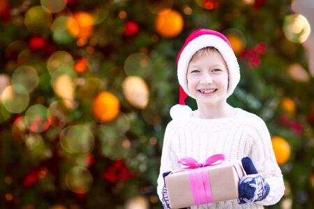 positive smiling child in santa's hat and warm winter clothes holding christmas gift enjoying christmas time by the treeの写真素材