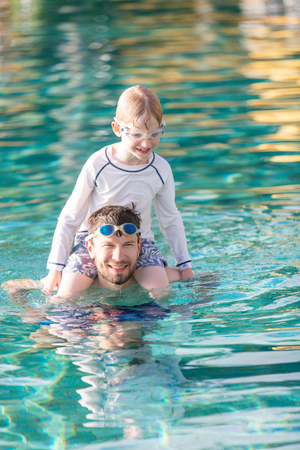 young father and smiling son enjoying and playing together in the pool, kid sitting on father shouldersの写真素材