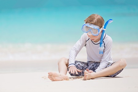 caucasian little boy in rashguard and with snorkeling kit sitting at picture perfect beach enjoying summer vacationの写真素材