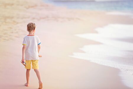 back view of little boy walking at the beautiful beach by the water at sunsetの写真素材