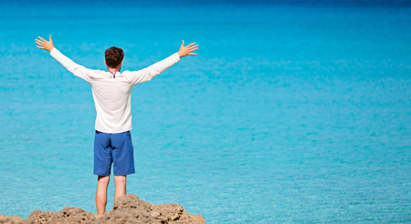 panorama of young man in rashguard and swimming shorts holding hands wide open and enjoying view of beautiful turquoise sea at perfect caribbean island, copyspace on rightの写真素材