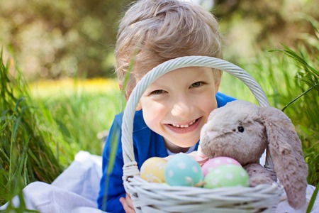 smiling caucasian boy after easter egg hunt with easter basket full of colorful eggs and bunny toy enjoying spring time in the parkの写真素材