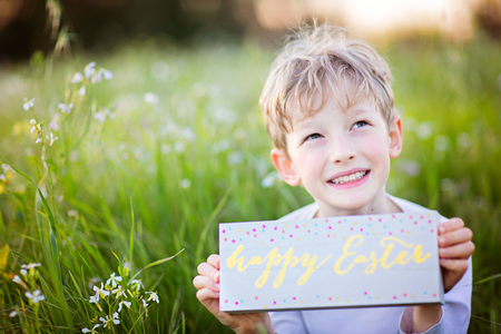 happy smiling boy holding colorful sign "happy easter" in hands enjoying beautiful blooming flowers in the parkの写真素材