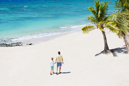 family of two, father and son, walking on empty beautiful white sand beach with palm trees and turquoise lagoon, family vacation concept, shot from aboveの写真素材