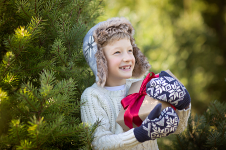 beautiful smiling boy in trapper hat and mittens holding christmas gift standing by the tree, winter holiday conceptの写真素材