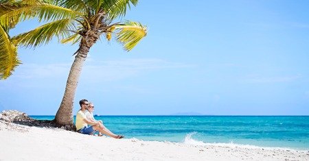panorama of beautiful family of two, father and son, sitting together at the beach by palm tree in the background enjoying summer vacation, white sand beach and turquoise lagoon, copy space on rightの写真素材