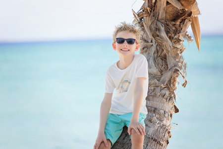 smiling positive boy in sunglasses standing by the palm tree at the beach, tropical vacation conceptの写真素材